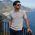 Homem usando Camisa Polo Italiana Ravello Masculina em tricô premium na cor cinza claro, óculos de sol e relógio, com fundo de paisagem litorânea europeia.
